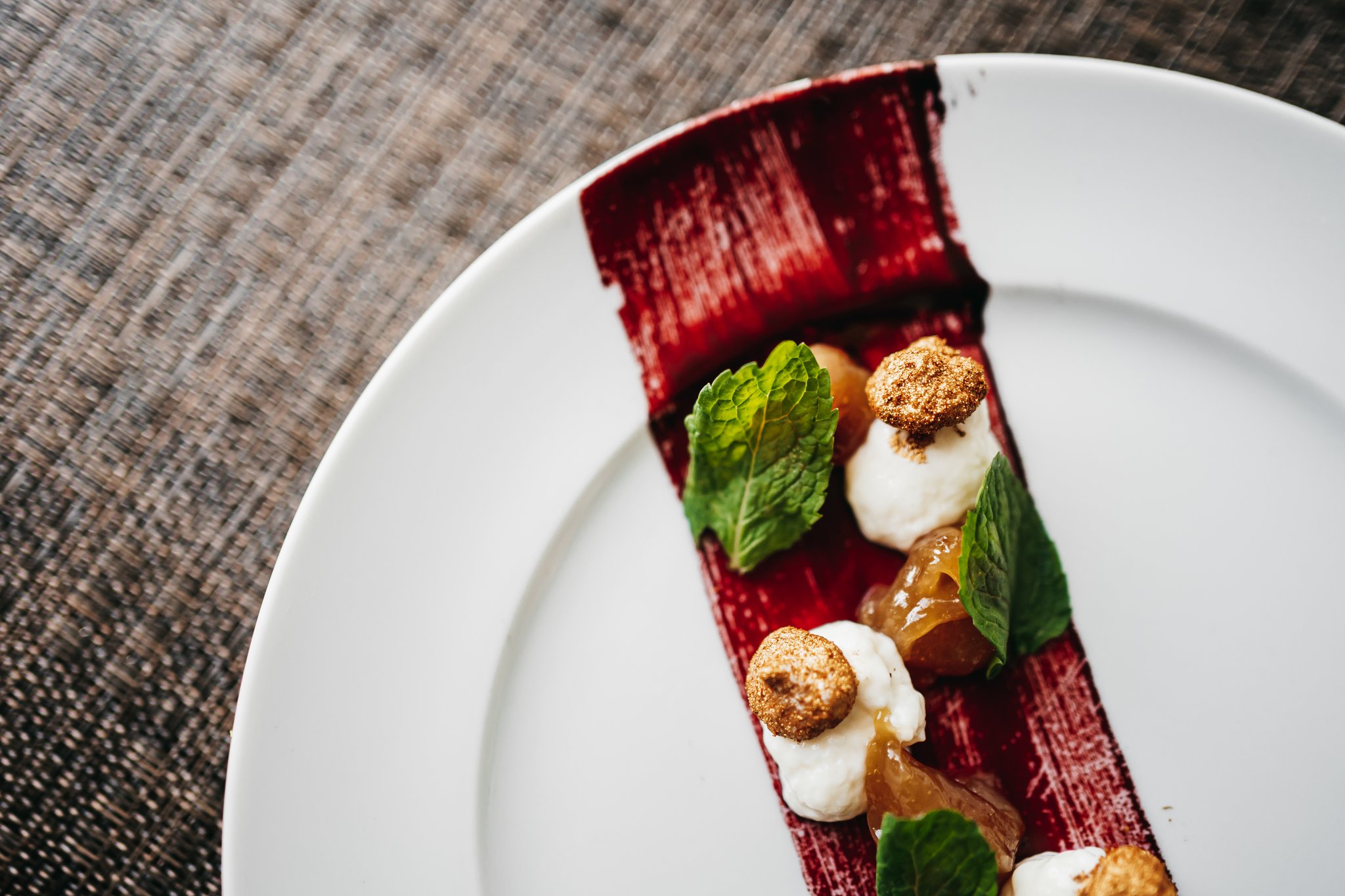 An elegantly plated dessert on a white round plate featuring a deep-red rectangular base topped with cream quenelles, spiced crumbs, translucent fruit gel, and fresh mint leaves, served aboard the untamed amazon cruise