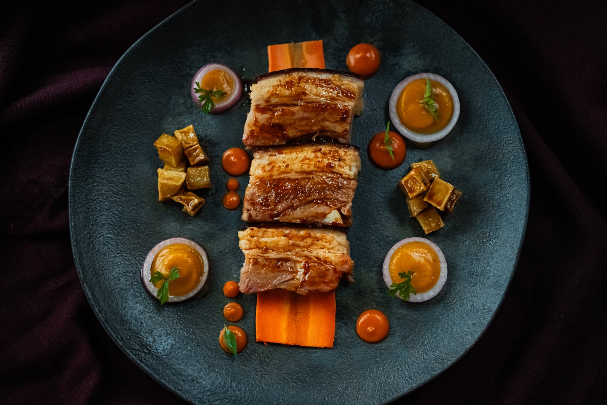 Three glazed slices of roasted meat rested on carrot ribbons, surrounded by golden sauce cups, cherry tomatoes and braised vegetable cubes on a teal plate — fine dining aboard the untamed amazon cruise