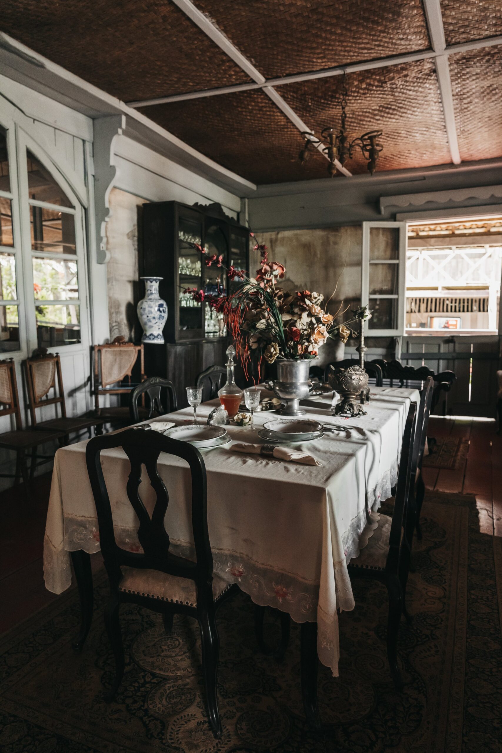 A formally set dining table with white linen, dark carved chairs, antique silverware, crystal glassware and a dried floral centrepiece inside a colonial-era room with a woven ceiling and vintage china cabinet — part of the untamed amazon cruise shore experience