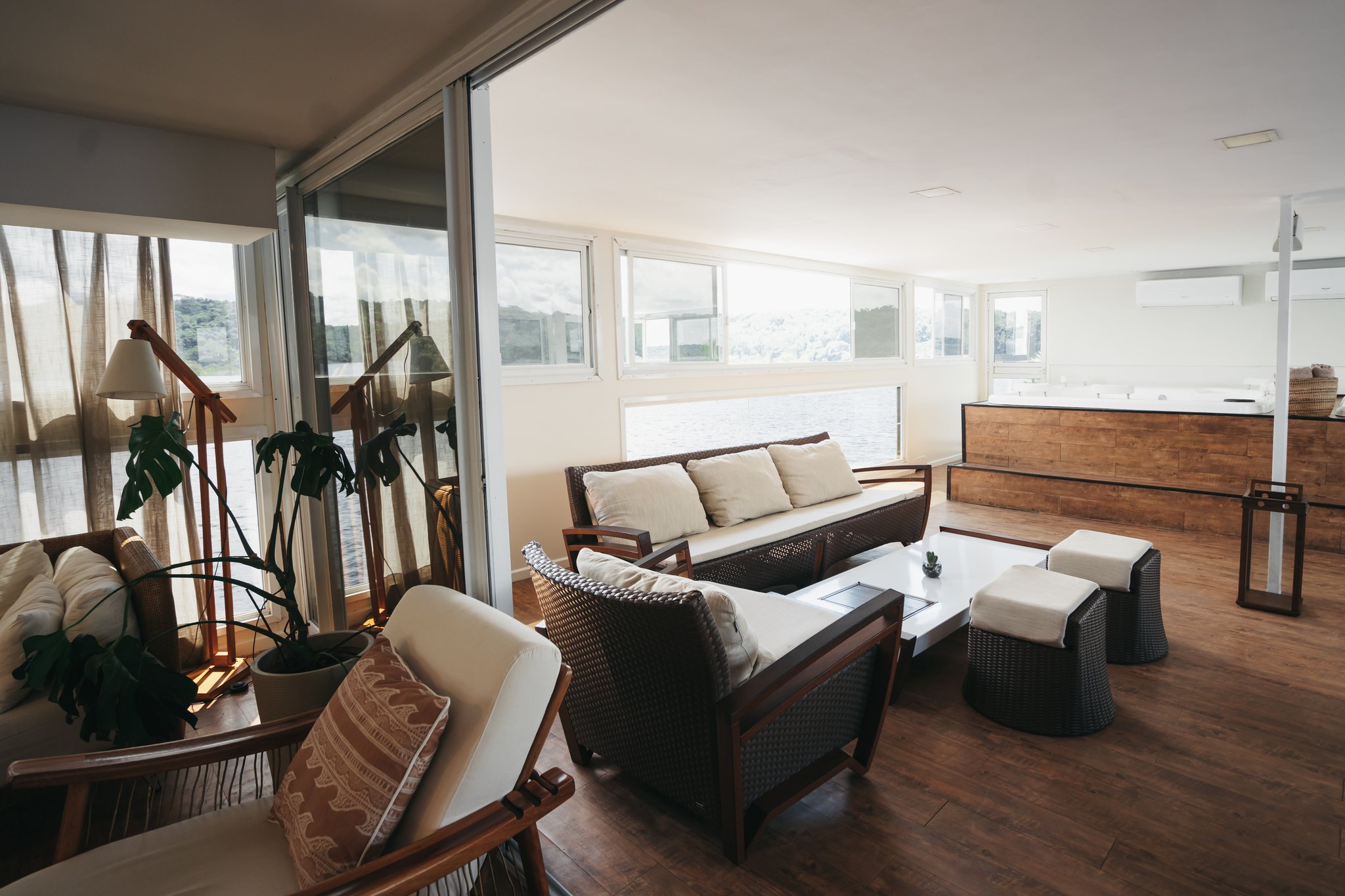 Bright, airy lounge on the untamed amazon cruise with rattan sofas, warm timber floors, a monstera plant, and panoramic windows overlooking the river