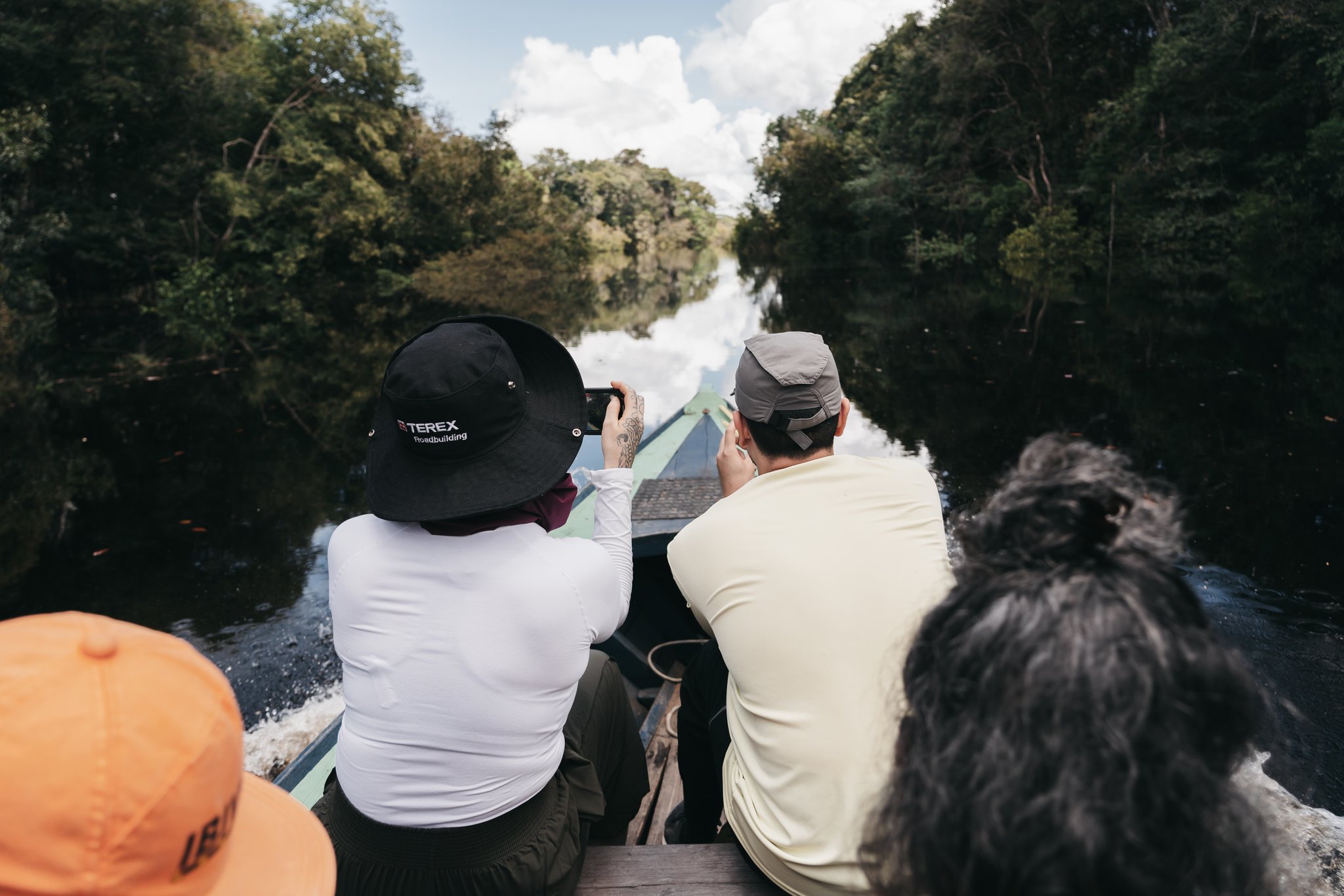 Ecotourists aboard a wooden boat photograph a mirror-still channel flanked by dense rainforest canopy on an untamed amazon cruise