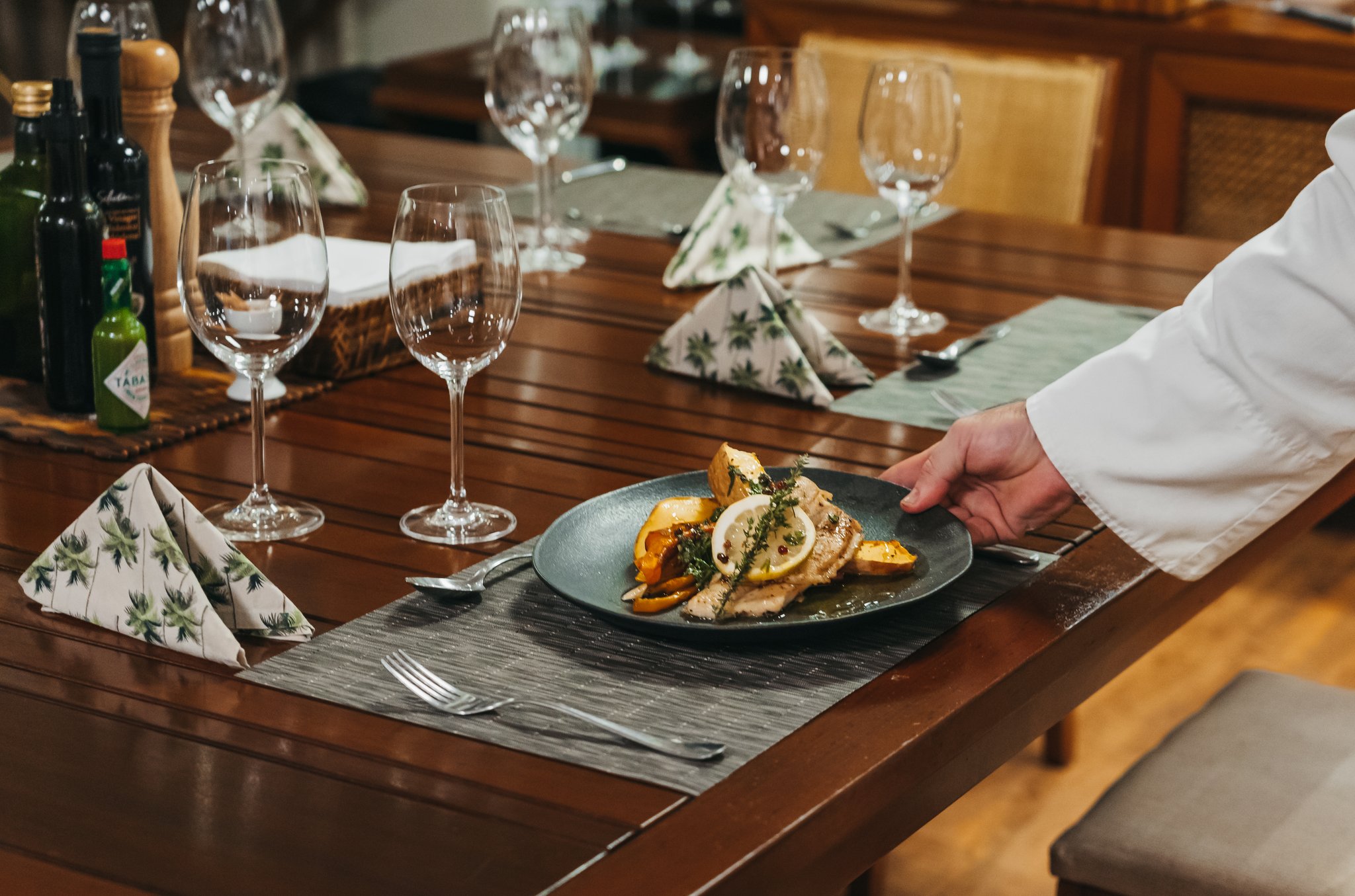 A chef in a white jacket placing a dark plate of grilled fish with roasted peppers, lemon and fresh herbs on a formally set wooden dining table aboard the untamed amazon cruise