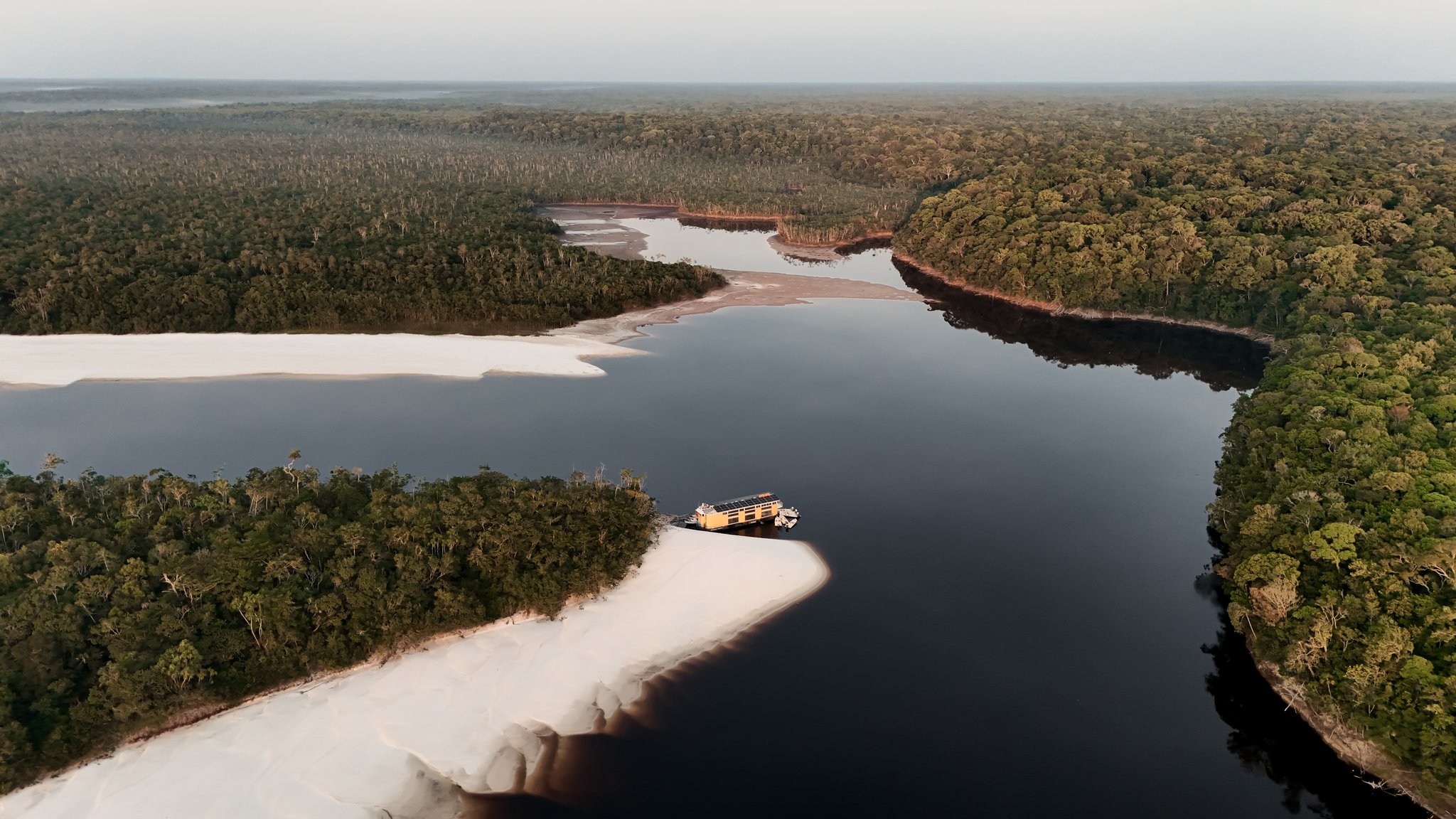Aerial view of a solar-powered expedition vessel moored beside a broad white sandbar on a dark-water Amazonian river, surrounded by dense rainforest canopy stretching to the horizon — an untamed Amazon cruise deep in the wilderness