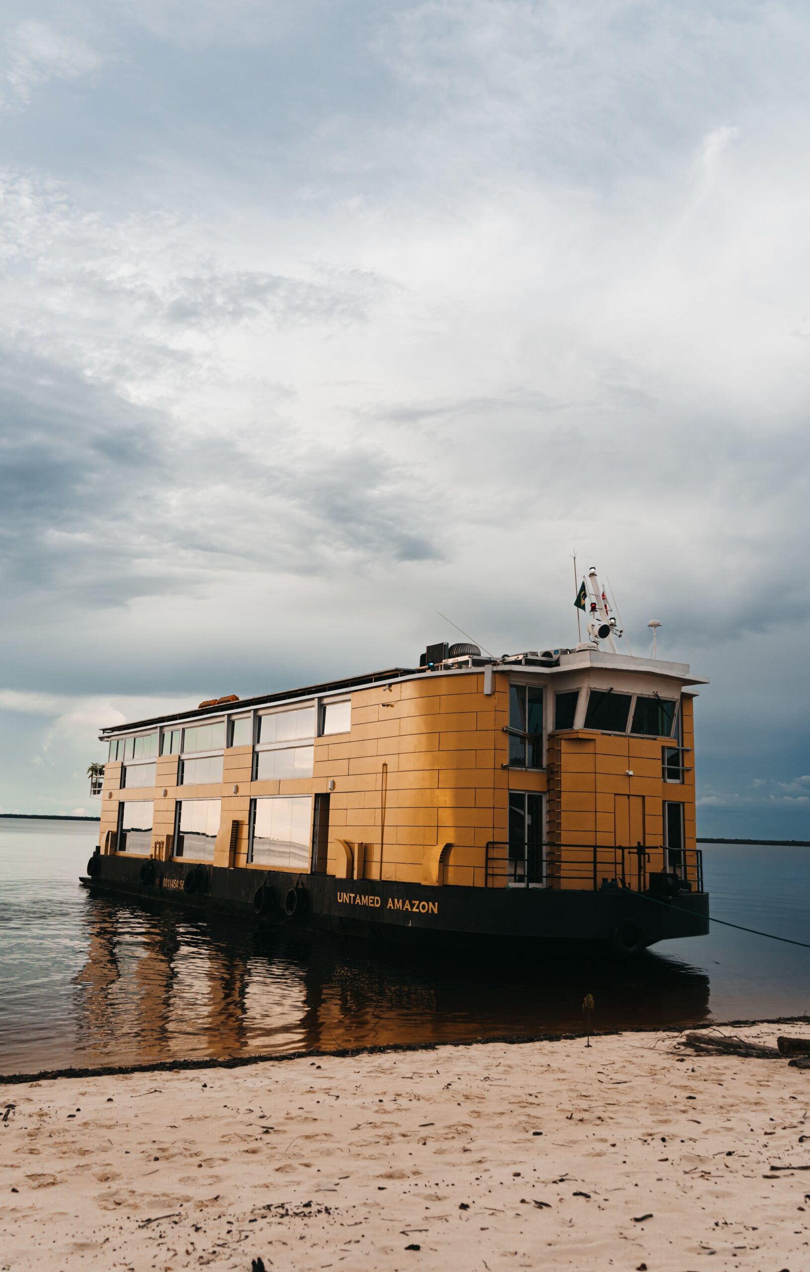 The Untamed Amazon cruise vessel, painted in golden yellow, moored beside a sandy Amazon riverbank under a dramatic cloudy sky