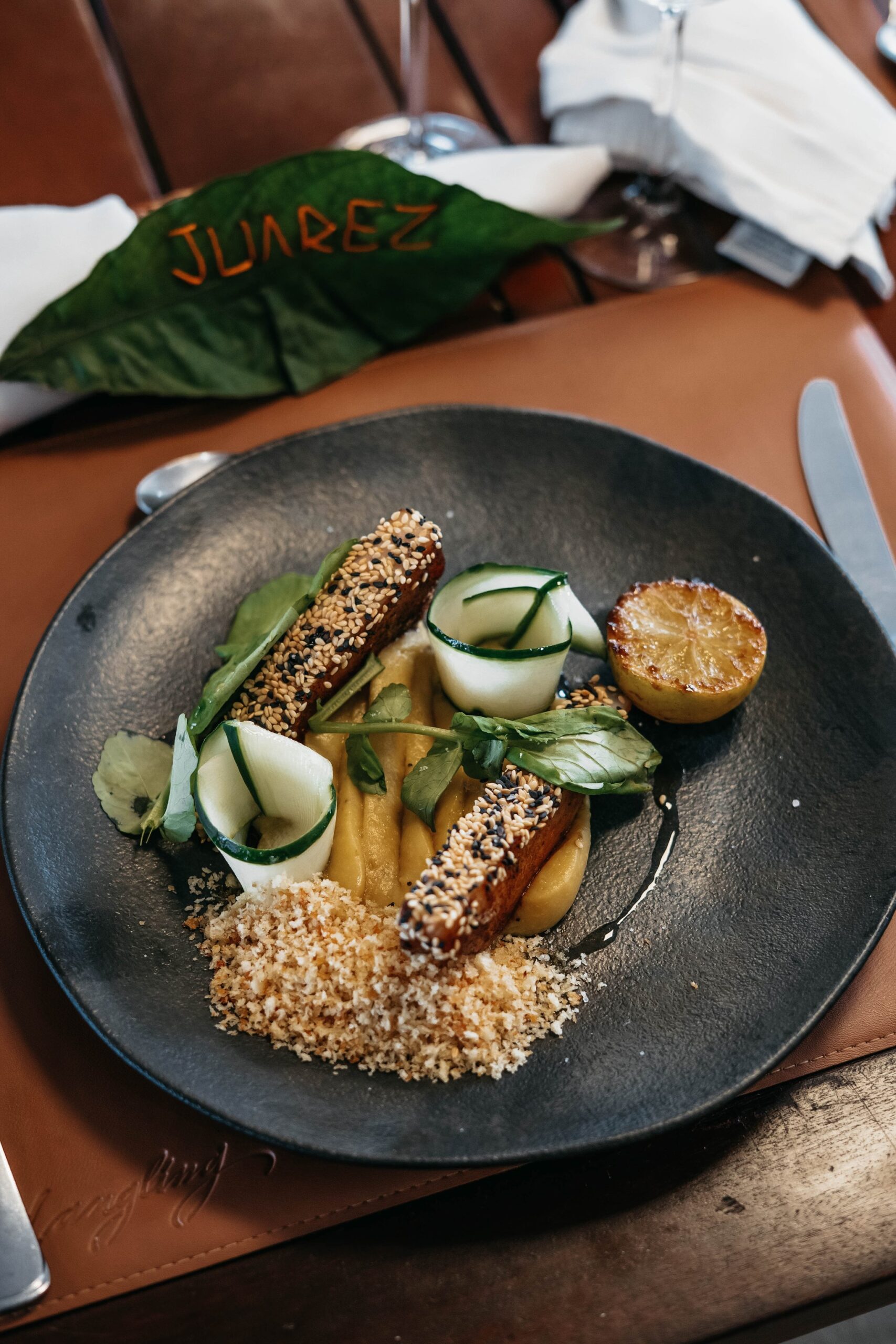 Sesame-crusted protein strips with cucumber rolls, golden purée, breadcrumbs and a charred citrus half plated on a dark slate dish aboard the untamed amazon cruise, with a leaf branded 'Juarez' in the background