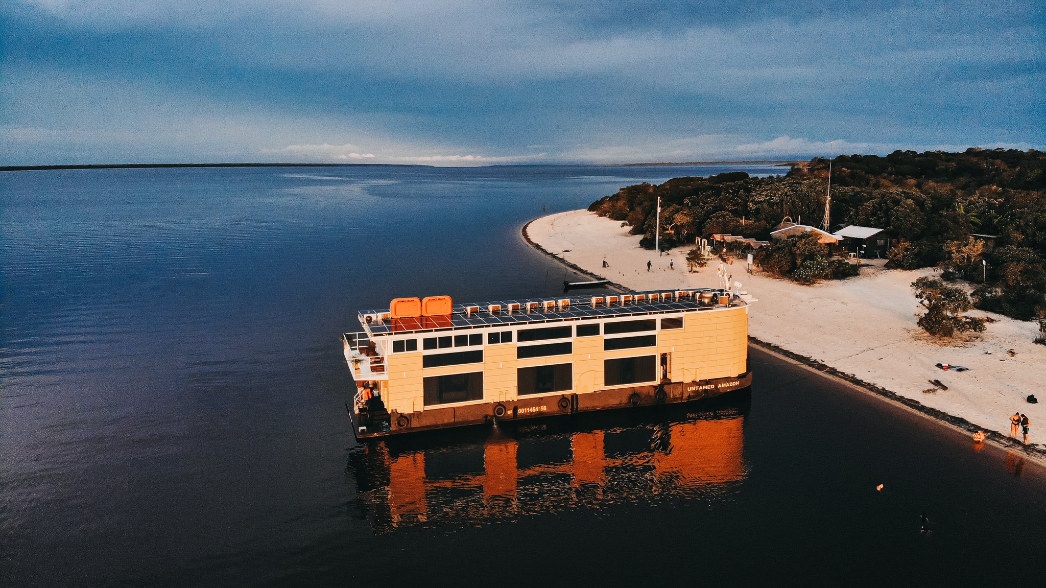 Aerial view of the Untamed Amazon Cruise solar-powered vessel moored beside a white-sand river beach at dusk, with dark water stretching to the horizon and forest-fringed shoreline behind