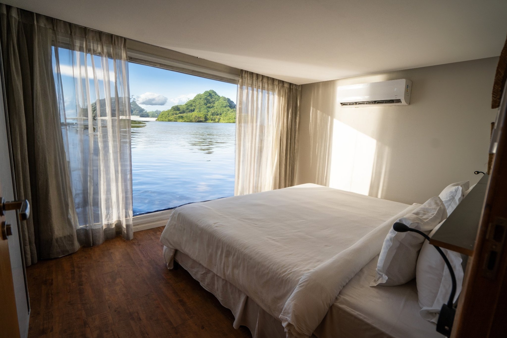 A bright, air-conditioned cabin on the untamed amazon cruise featuring a double bed with white linen, hardwood floors, and a floor-to-ceiling window overlooking a calm river and tree-covered island