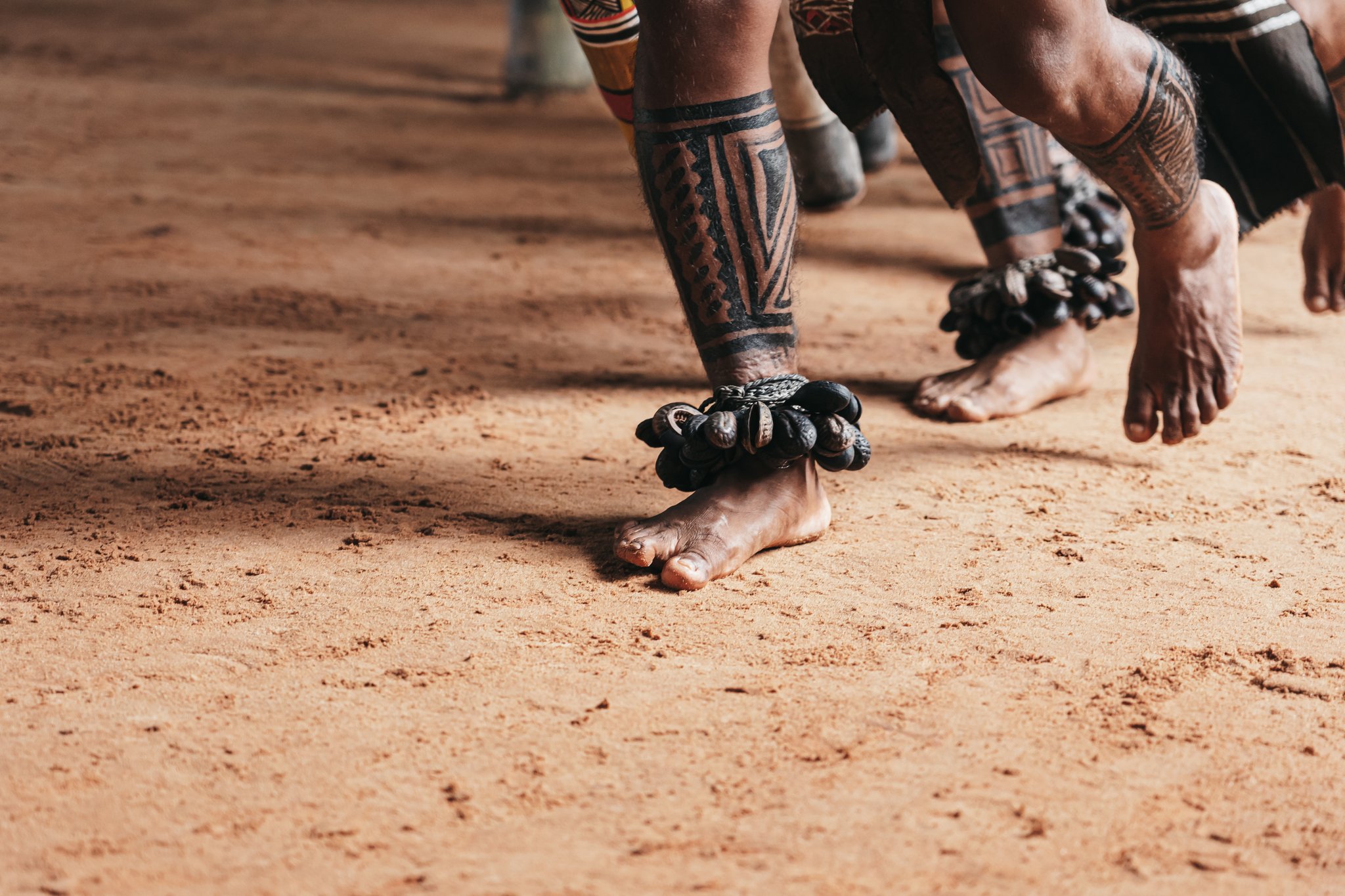 Close-up of bare feet on red sand earth, adorned with seed anklets and bold geometric body paint on the legs of indigenous dancers during an untamed amazon cruise community visit