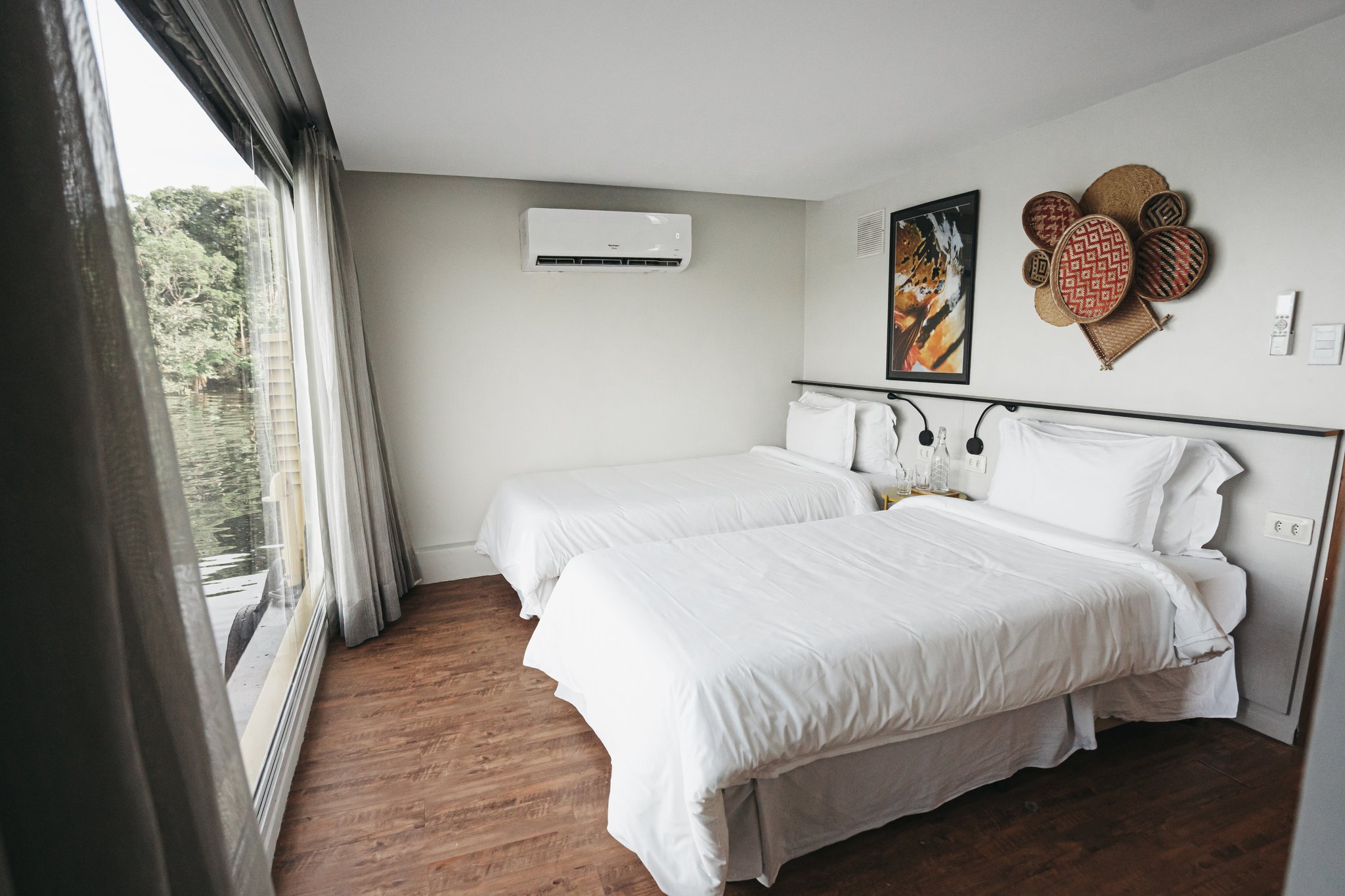 Twin-bed cabin on the untamed amazon cruise with crisp white bedding, hardwood floors, woven basket wall decor, and floor-to-ceiling windows overlooking a green riverbank