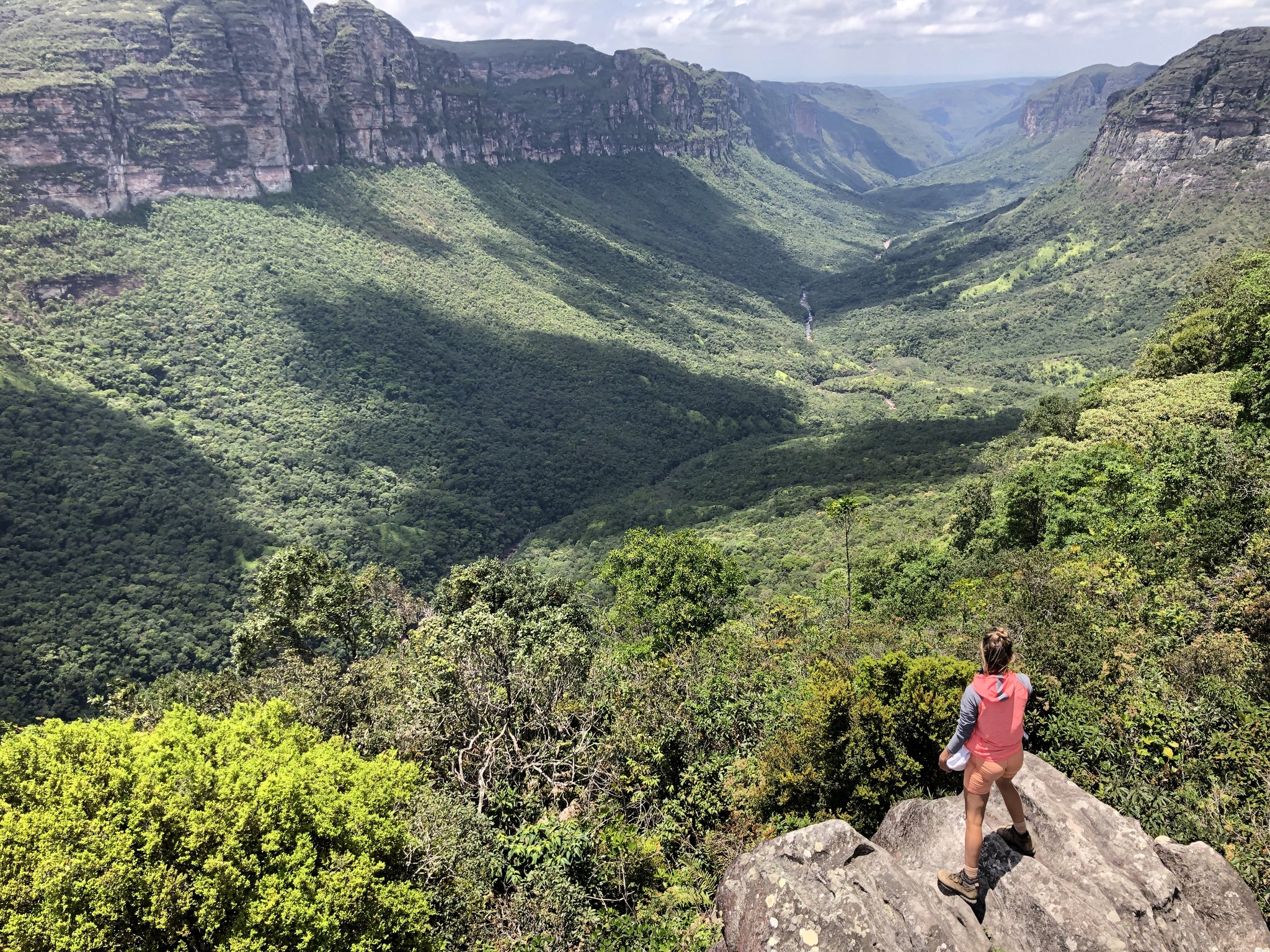 A canyon so wide it swallows your sense of scale. Photo: Lucas Neves A hiker stands on a rocky outcrop overlooking the vast green canyon of the Vale do Pati Trek, with towering sandstone escarpments and a distant waterfall visible deep in the valley