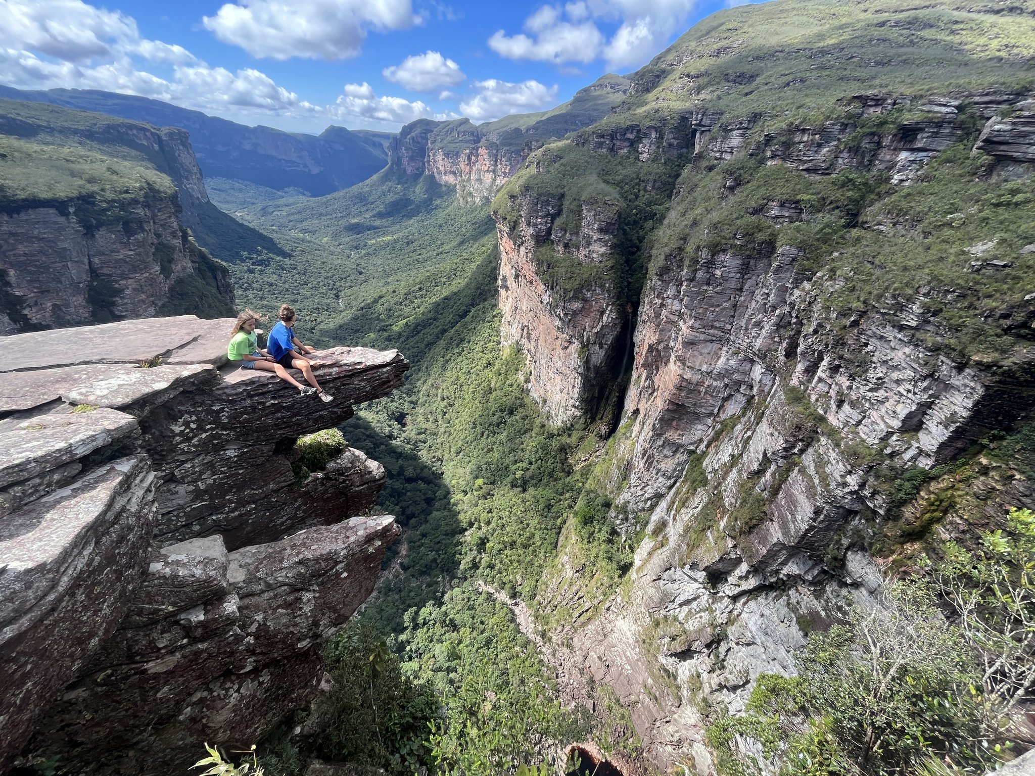 Legs dangling, abyss below — the canyon earns every uphill step. Two hikers sitting on a rock ledge overlooking the deep canyon and lush green valley of the Vale do Pati Trek in Chapada Diamantina