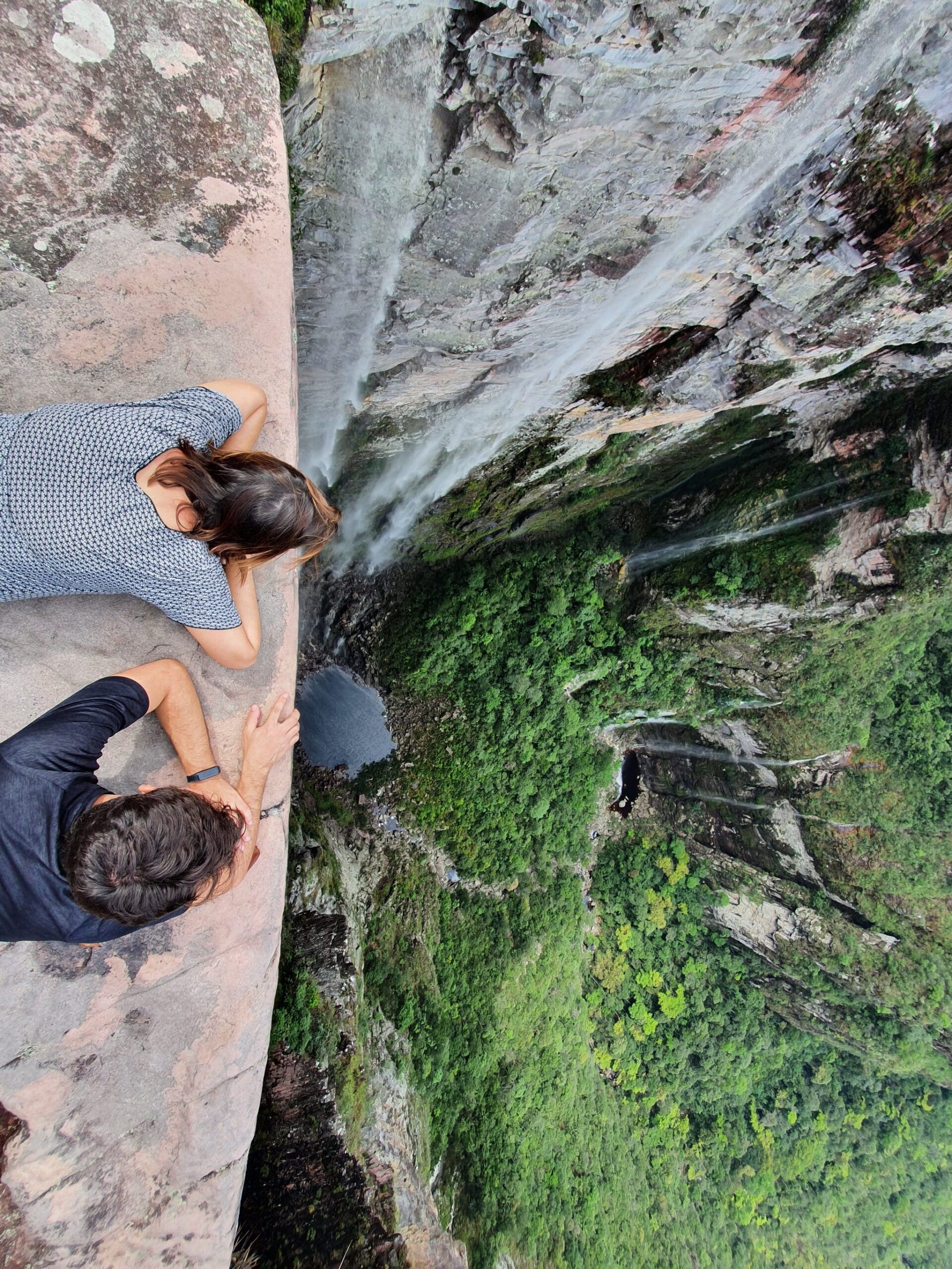 Stomach to the rock, eyes on the abyss — this is what 260 metres of air feels like. Photo: Lucas Neves Two hikers lie flat on a smooth sandstone ledge peering straight down a towering cliff face on the Vale do Pati Trek, with a thin waterfall and dense green canyon far below