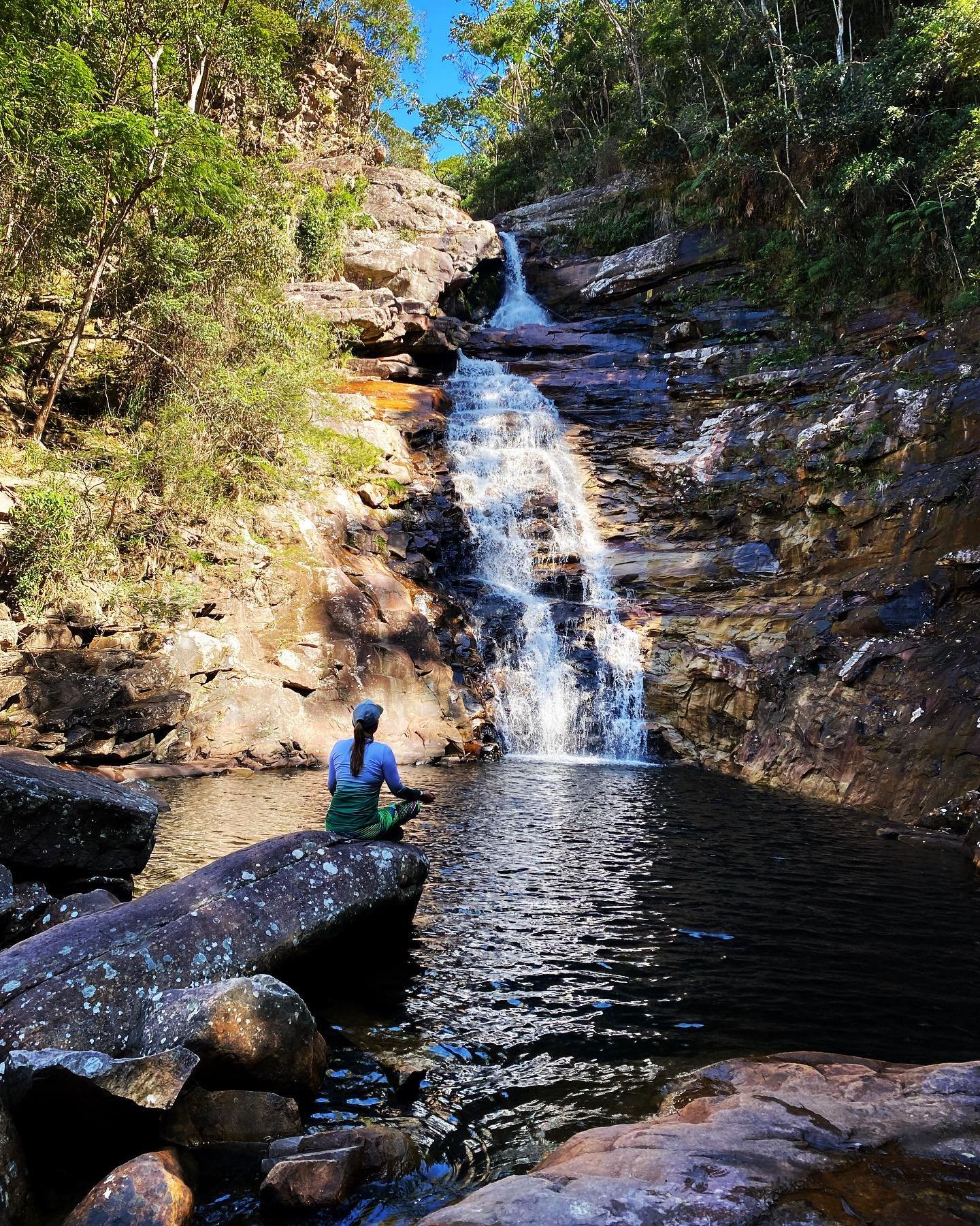 Still water, rushing falls — a well-earned rest at the pool's edge. A hiker sits on a rock beside the natural pool of a tiered waterfall cascading down iron-streaked sandstone cliffs on the Vale do Pati Trek, surrounded by green Atlantic forest under a blue sky