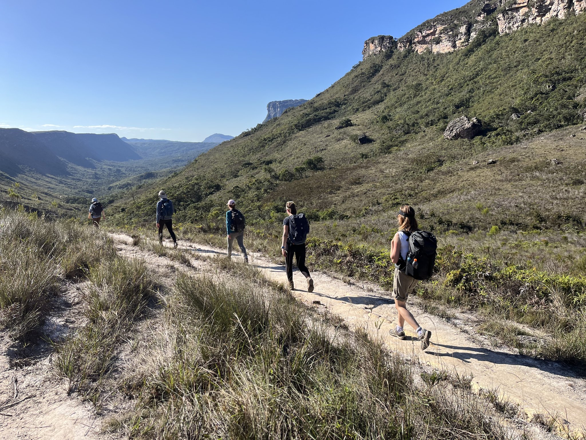 Five hikers, one trail, a valley that keeps opening up. Five backpackers hiking single-file along a sandy stone trail on the Vale do Pati Trek, with sweeping valley views and towering sandstone escarpments under a clear blue sky