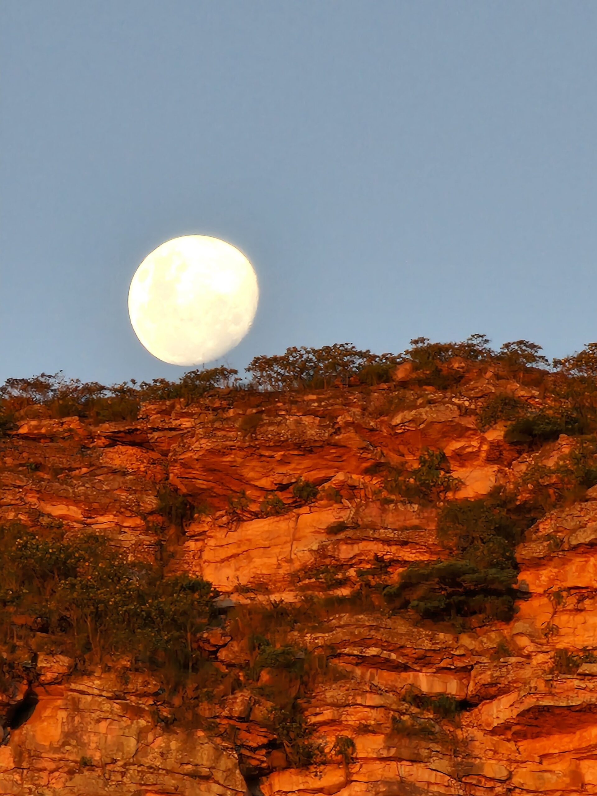 Moon up, cliffs still burning — the valley keeps its own time. A near-full moon rising above warm red sandstone cliffs dotted with scrub vegetation on the Vale do Pati Trek, under a clear blue twilight sky
