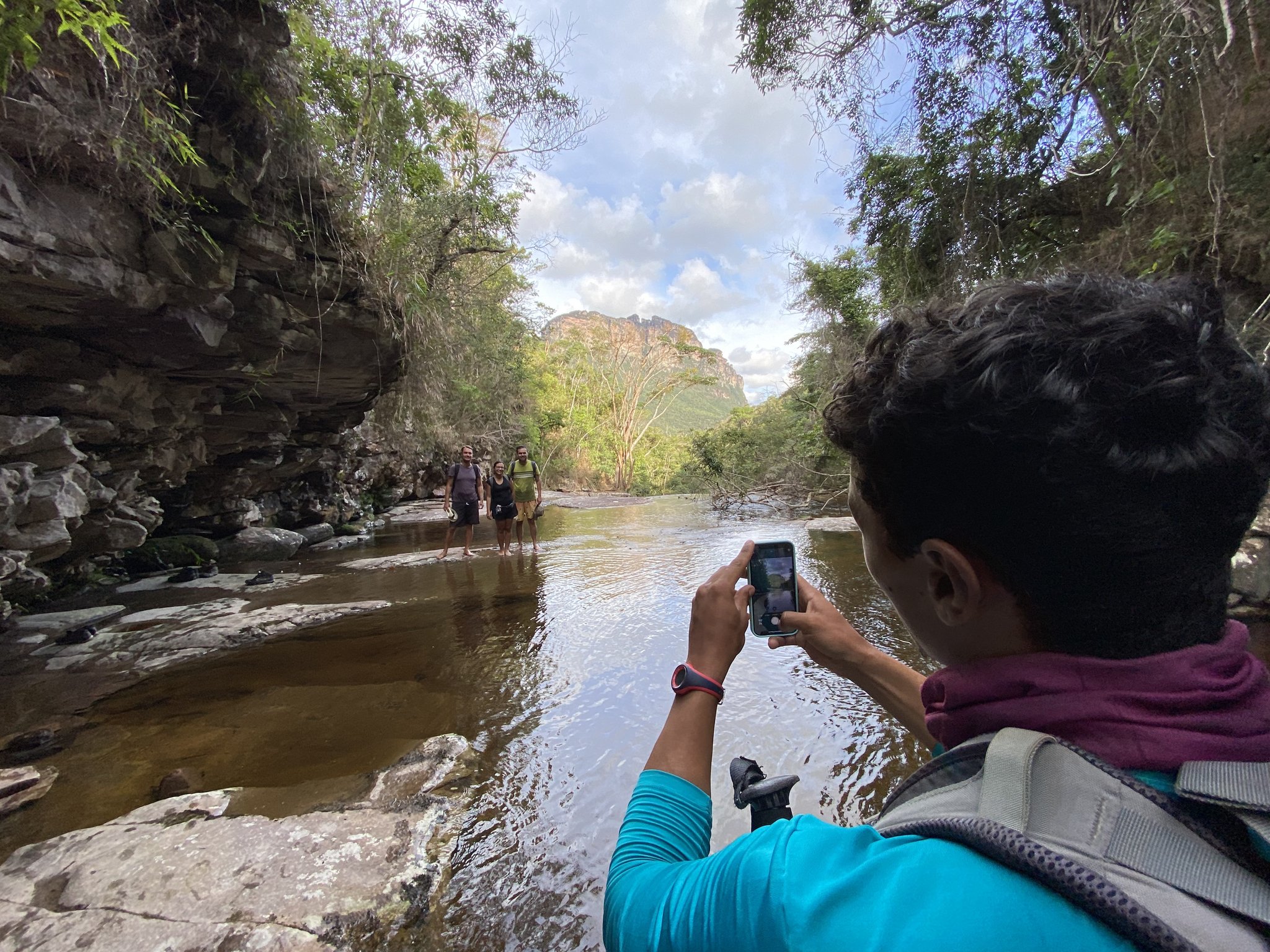 Phone out, friends across the water — memories made mid-stream. A hiker photographs two smiling trekkers standing in a shallow rocky river gorge on the Vale do Pati Trek, with sandstone cliffs and a mesa visible in the background