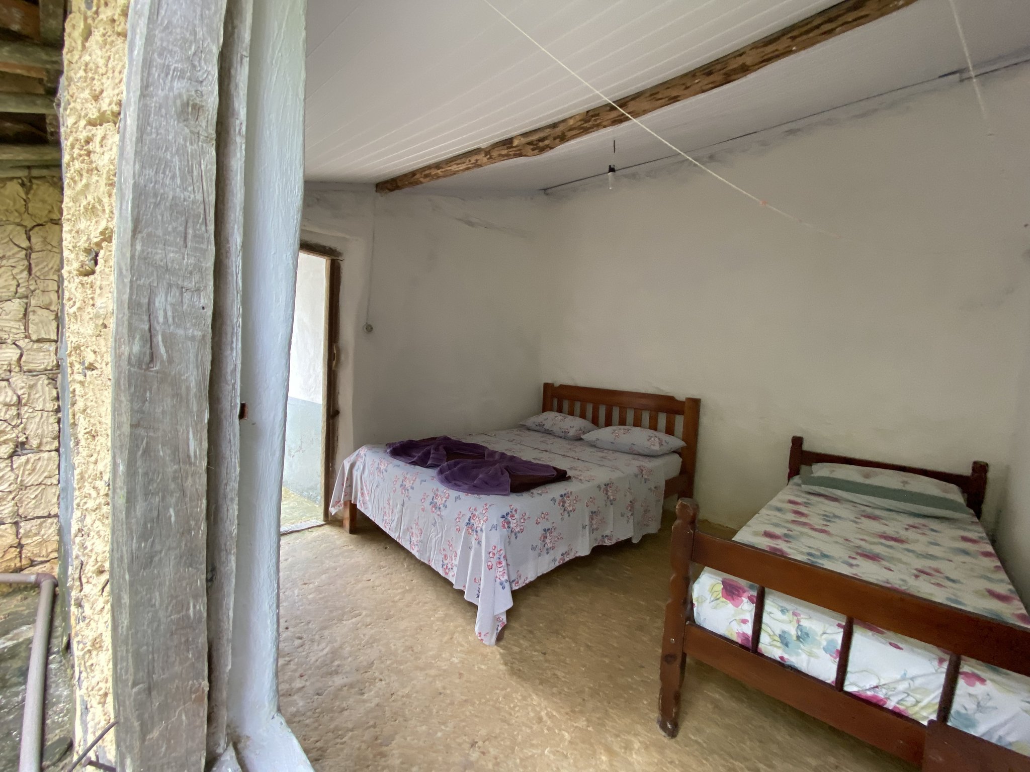 Rustic beds, stone walls, and a door open to mountain air — simple comforts deep in the valley. Simple shared bedroom in a Vale do Pati Trek family homestay, with two wooden-framed beds covered in floral bedspreads, exposed ceiling beam, and stone-and-plaster walls