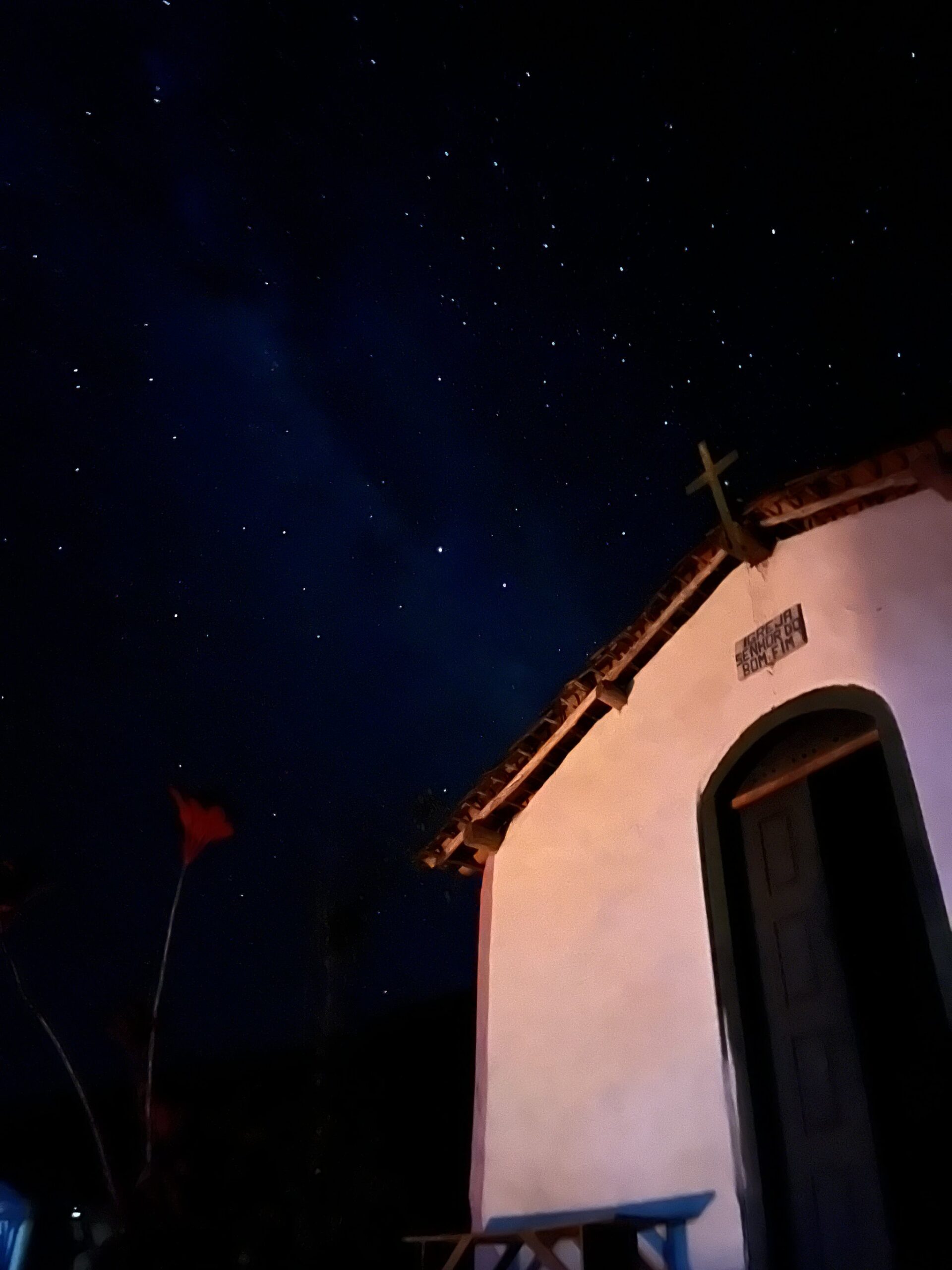 Stars thick overhead, a tiny chapel below — the valley sleeps. A whitewashed colonial chapel signed 'Igreja Senhor do Bonfim' beneath a brilliant starry sky on the Vale do Pati Trek, with a wooden cross silhouetted against the deep blue night