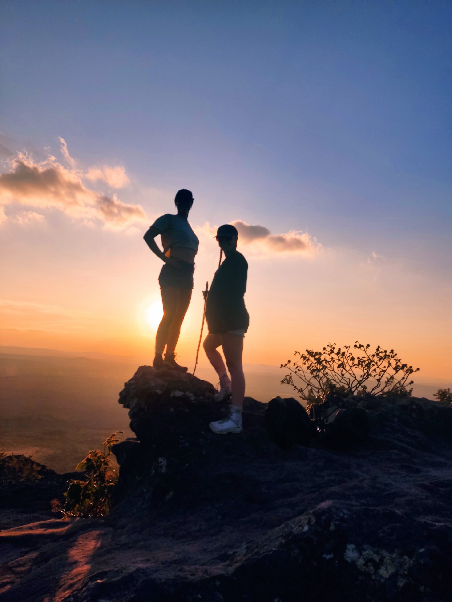 Two hikers silhouetted against a golden sunset on a rocky summit during the vale do pati trek, with an expansive valley stretching to the horizon below