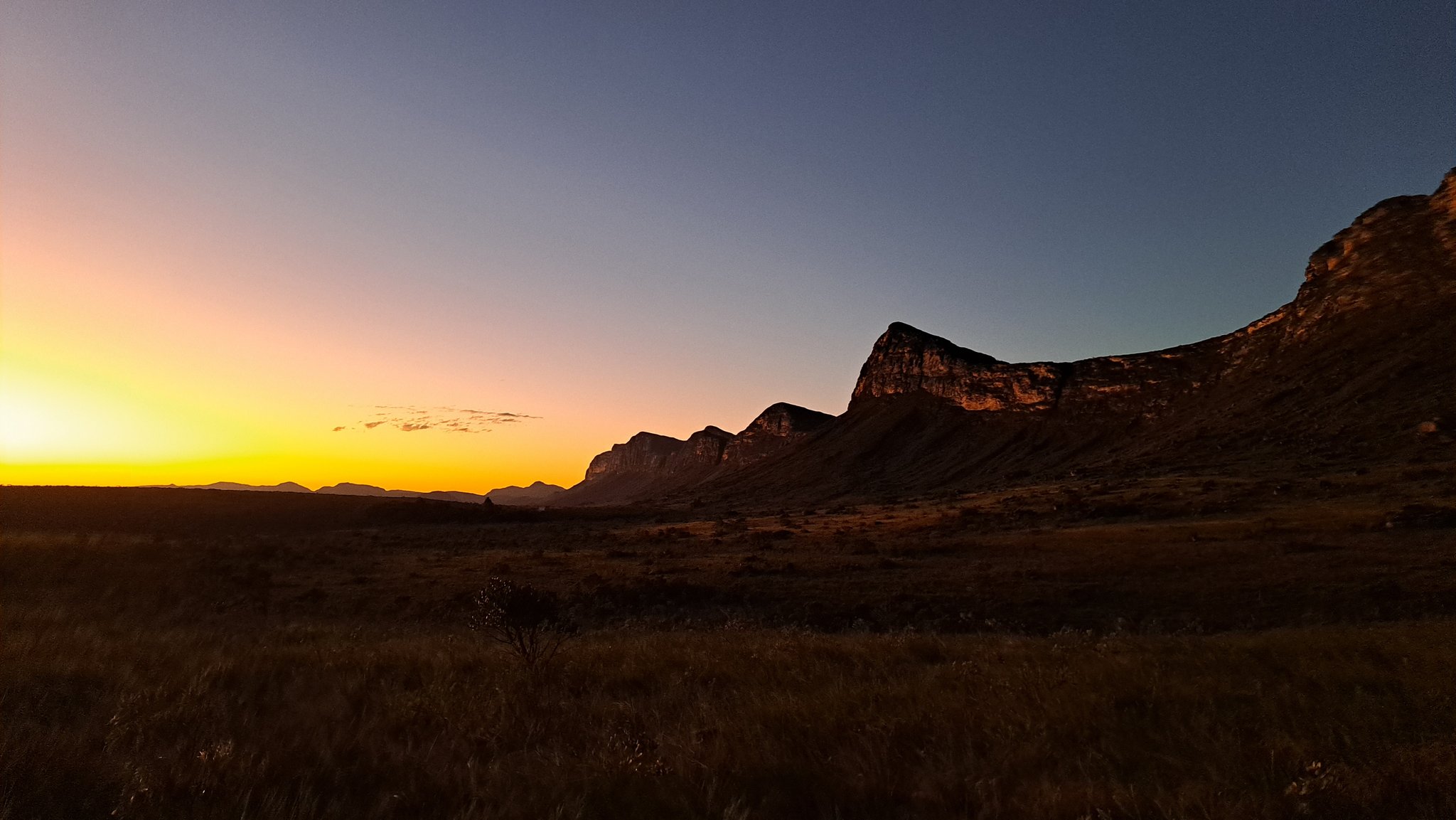 Last light bleeds gold across the plains, the escarpment holding its shape against the darkening sky. Fiery orange and gold sunset over a vast open savanna with a rugged rocky escarpment silhouetted against a deep blue sky on the Vale do Pati Trek