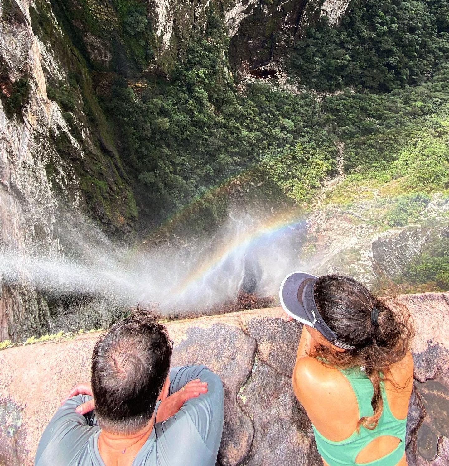 Two hikers crouch at the rocky edge of a towering waterfall on the vale do pati trek, mist filling the air and a vivid rainbow forming in the spray above the jungle canyon below