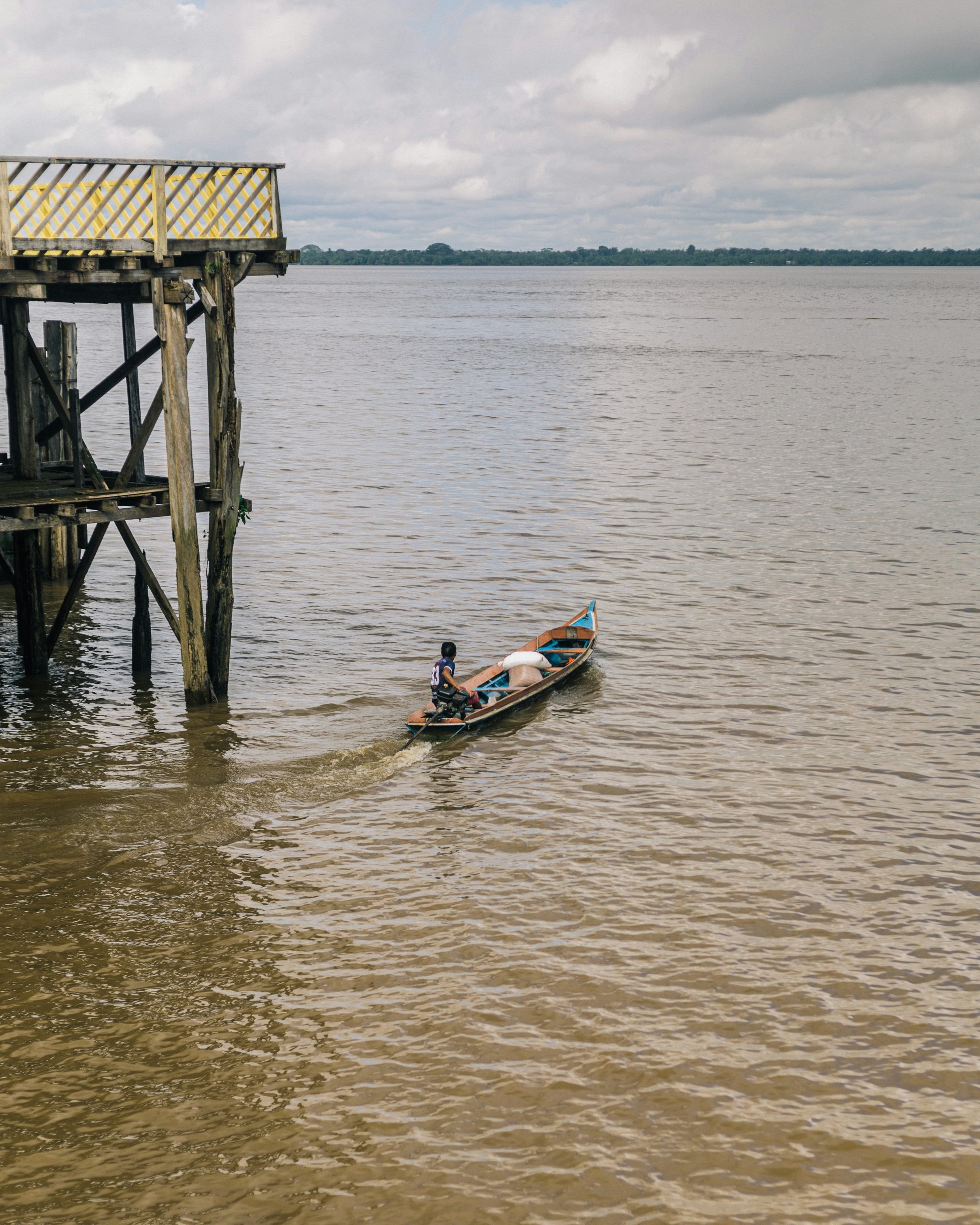Small motorized canoe passing a wooden stilt pier on the wide muddy Amazon river near Manaus