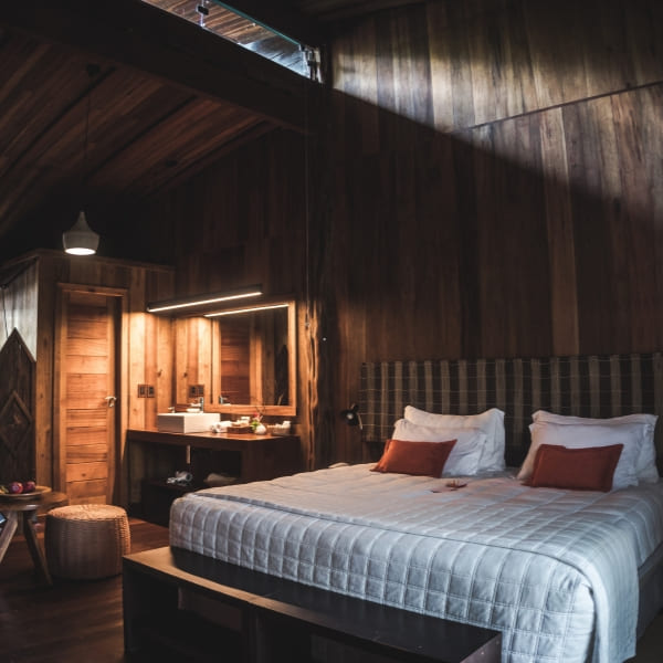 A wood-panelled luxury suite bedroom with a large bed, orange cushions and a vanity, warmly lit, at an Amazon lodge.