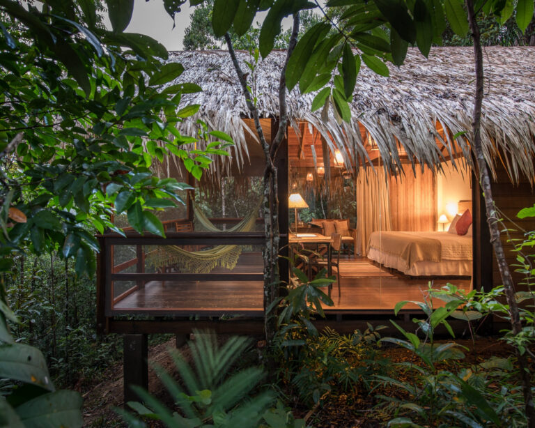 A luxury Amazon jungle lodge bungalow with a thatched roof and glass walls glowing with warm light at dusk, a hammock on its wooden deck amid the rainforest.